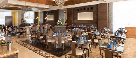 A spacious hotel lobby with multiple dining tables and chairs, a large chandelier, and a Christmas tree.