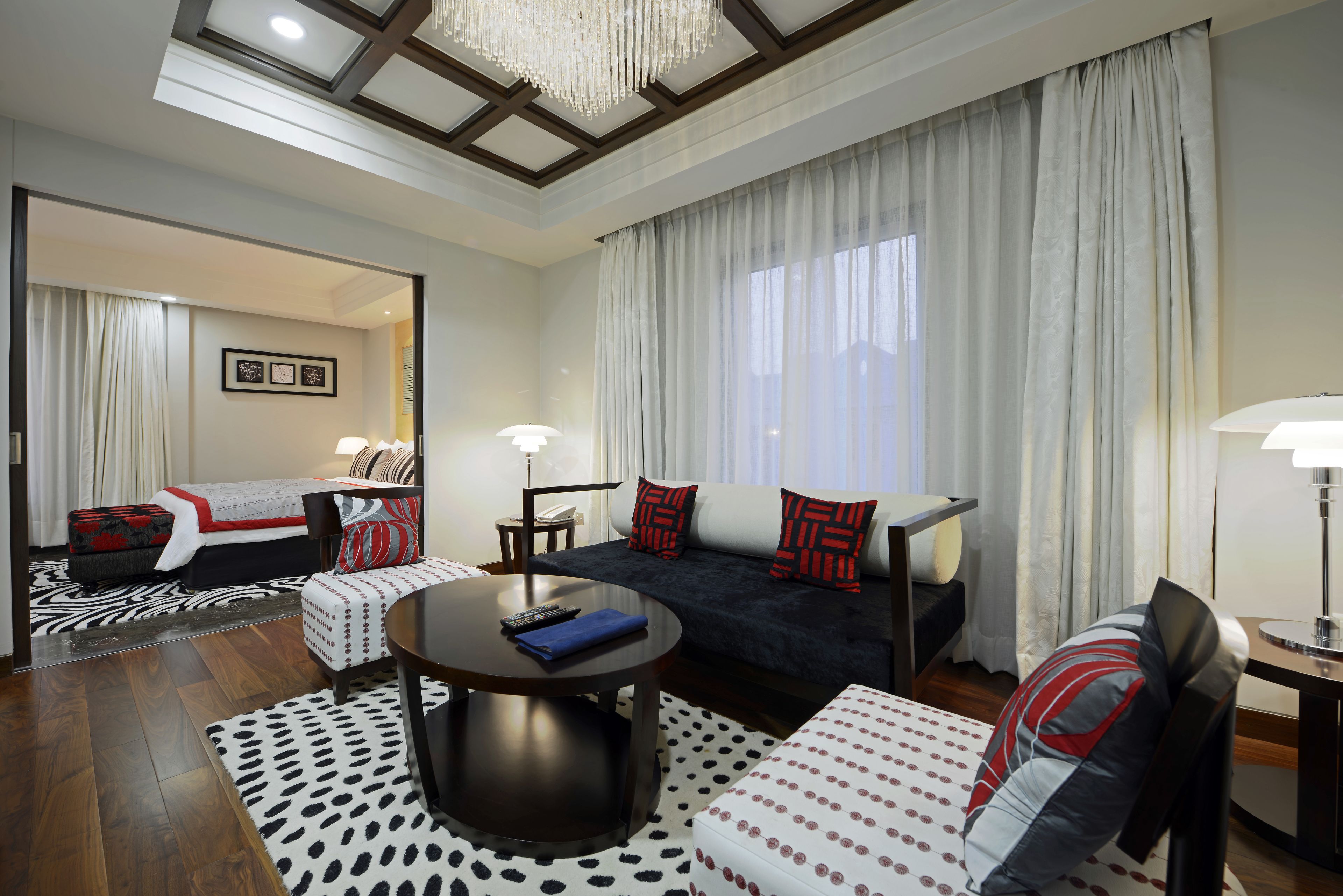A modern hotel room with a sofa, armchair, and a round table. There is a bed with a red and black patterned bedspread, and a large window with sheer curtains.