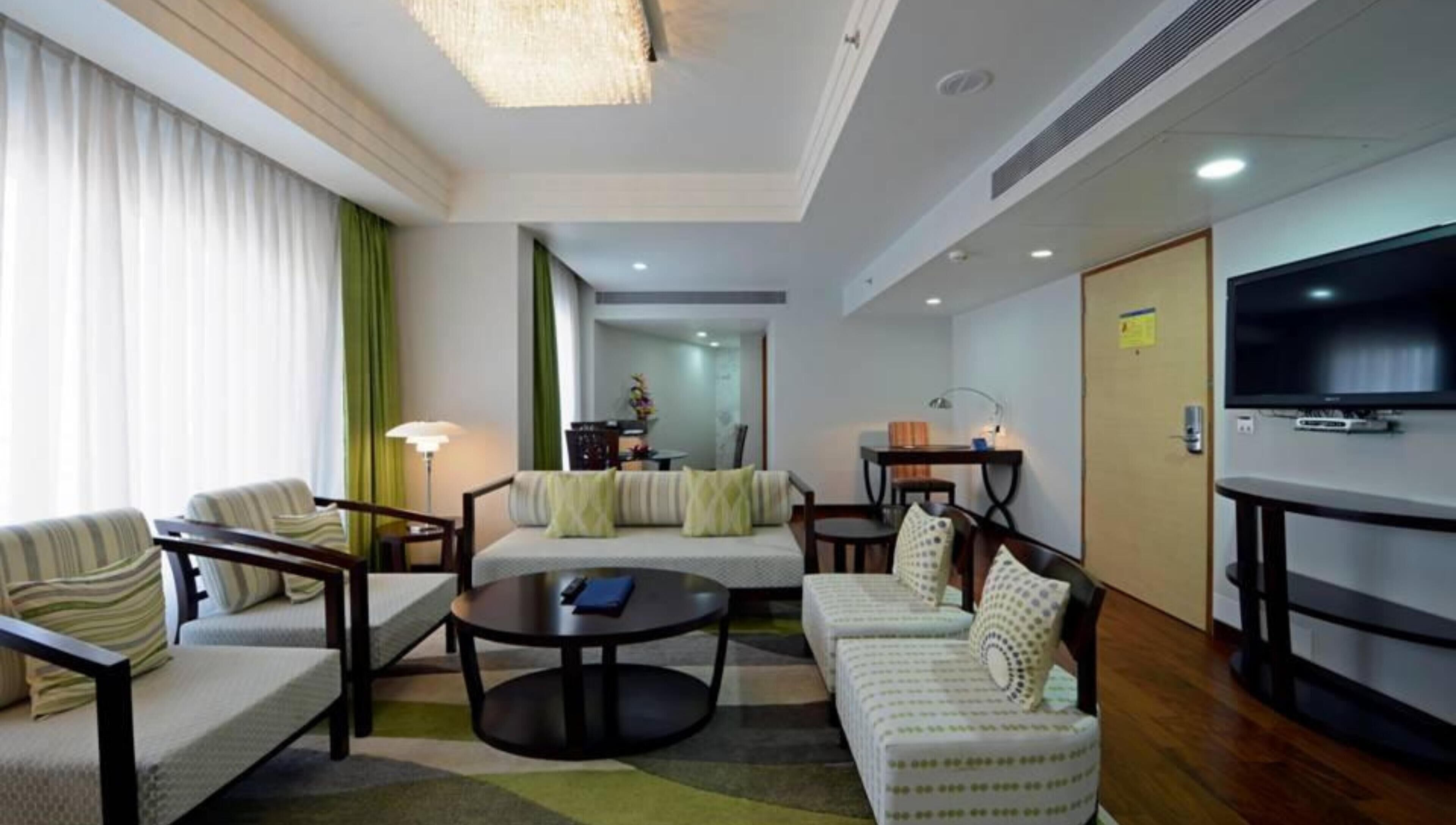 A modern hotel room with a sofa, armchairs, a coffee table, a TV, and a dining area.