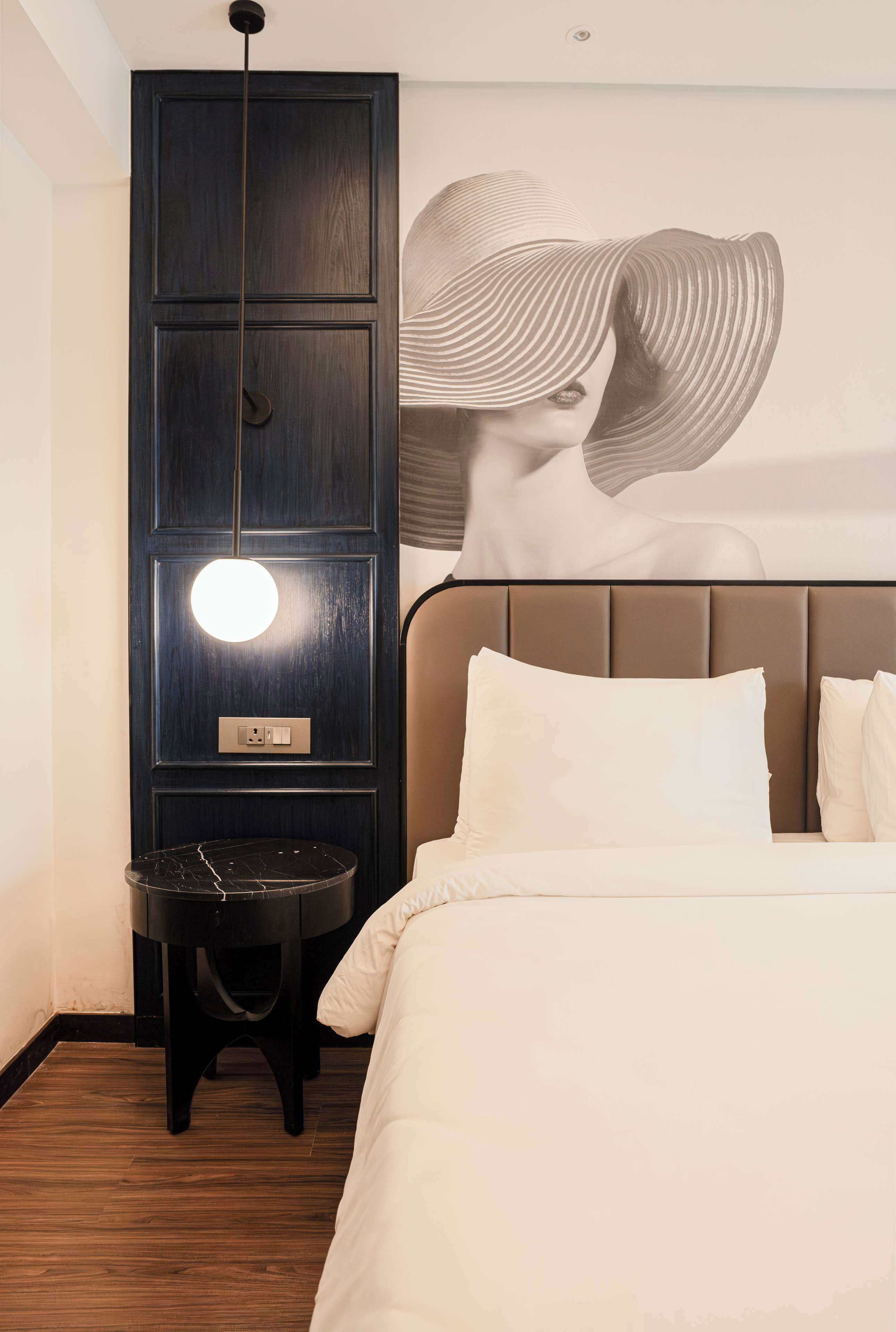 A hotel room with a bed, a round table, a wall-mounted light, and a large abstract artwork.