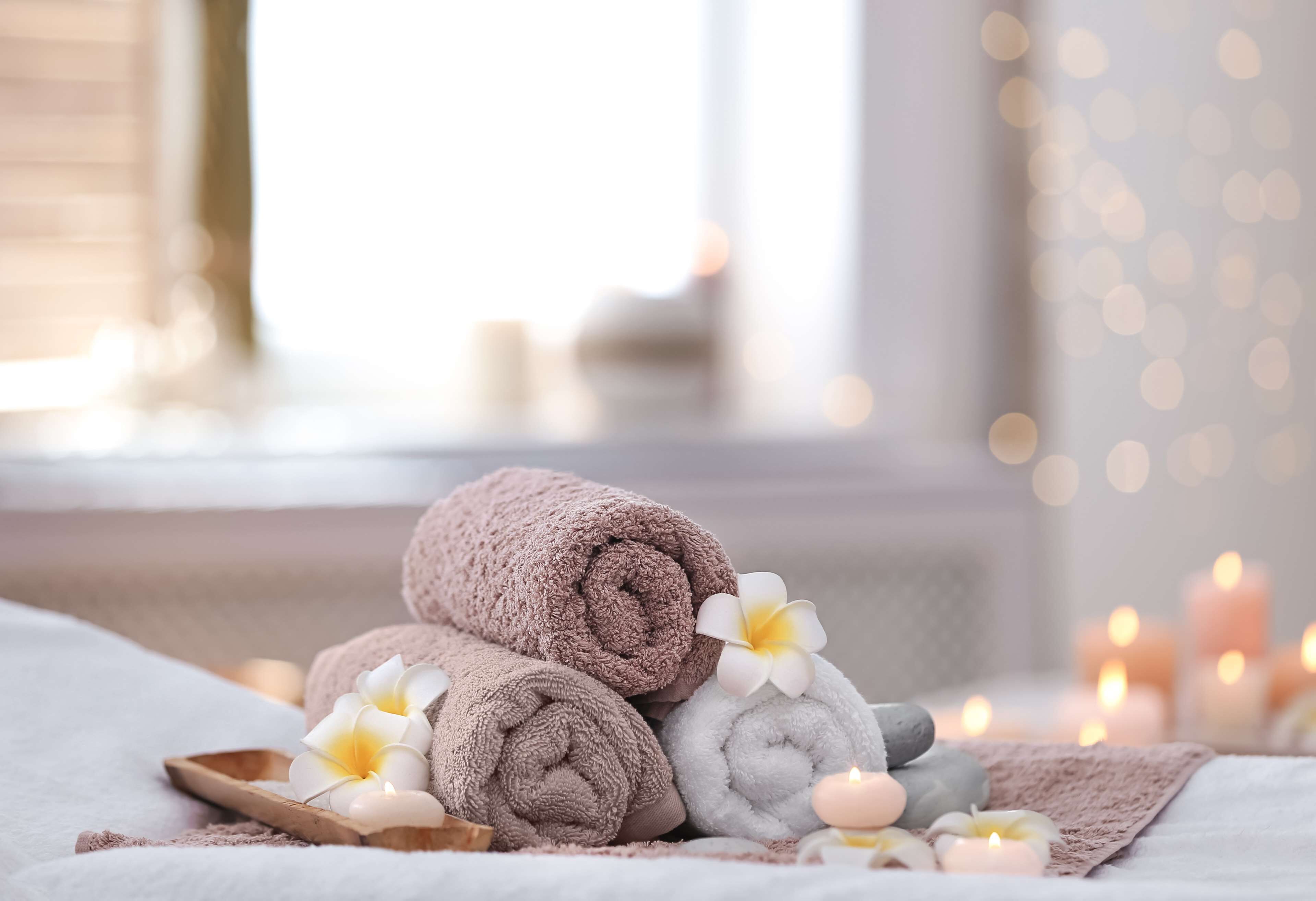 A neatly arranged stack of towels and a wooden tray with a flower, set against a softly lit background with candles.