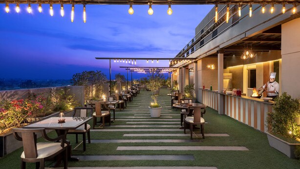 Un espacio exterior para comer con mesas y sillas, un chef preparando comida y vistas de la ciudad al atardecer.