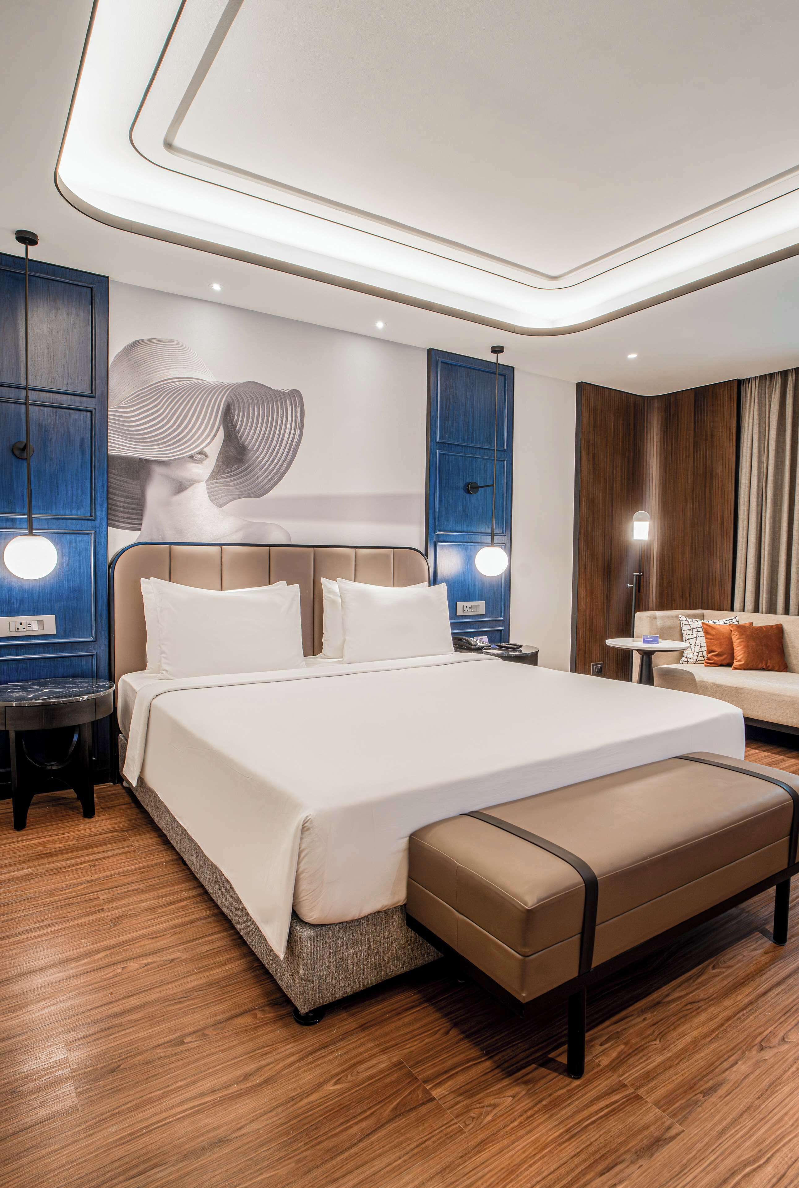 A modern hotel room with a large bed, a bench, a sofa, and a wall mural.