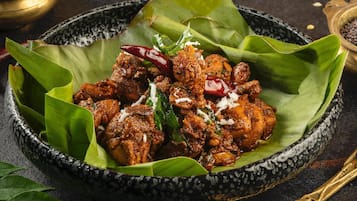 Un tazón de curry de pollo con hoja de plátano sobre una superficie oscura, acompañado de un tazón de pan naan y una cuchara con semillas.