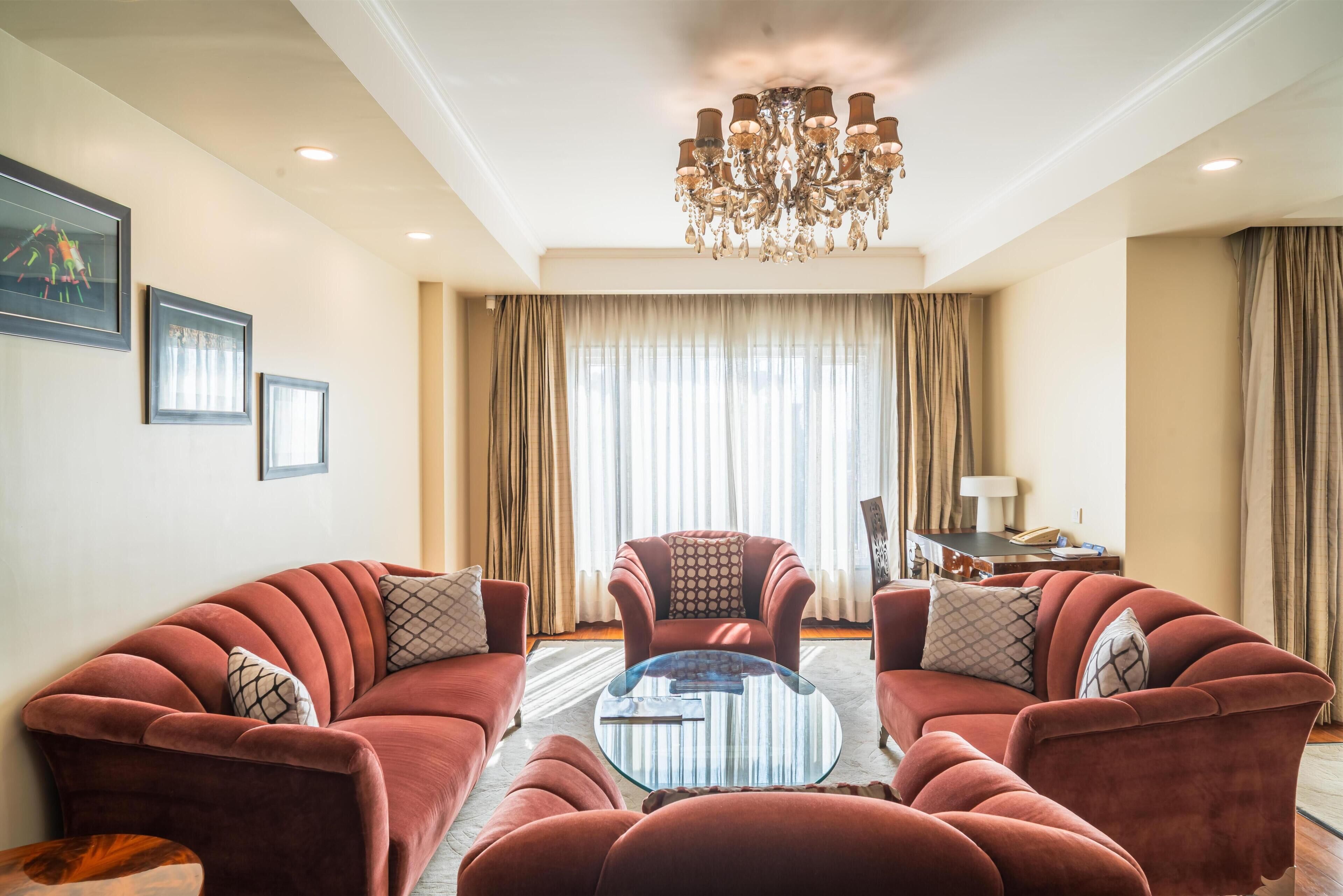 A living room with a red sofa set, a glass coffee table, a chandelier, and a large window with curtains.