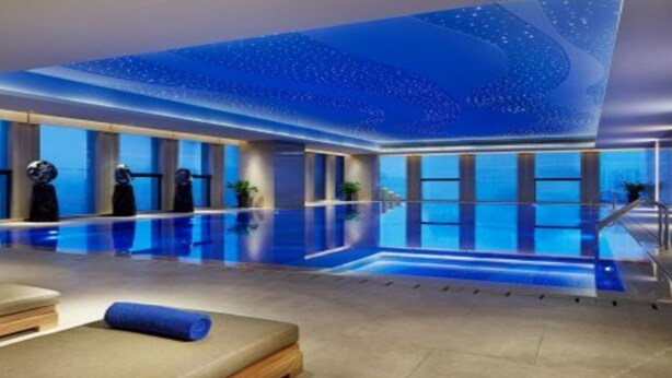 Un área de piscina interior moderna con superficie reflectante, sillones de descanso y techo con un diseño luminoso.