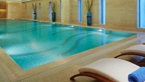 En indendørs swimmingpool med klart blåt vand, omgivet af beige vægge og vinduer. Liggestole med håndklæder og dekorative planter langs væggene.