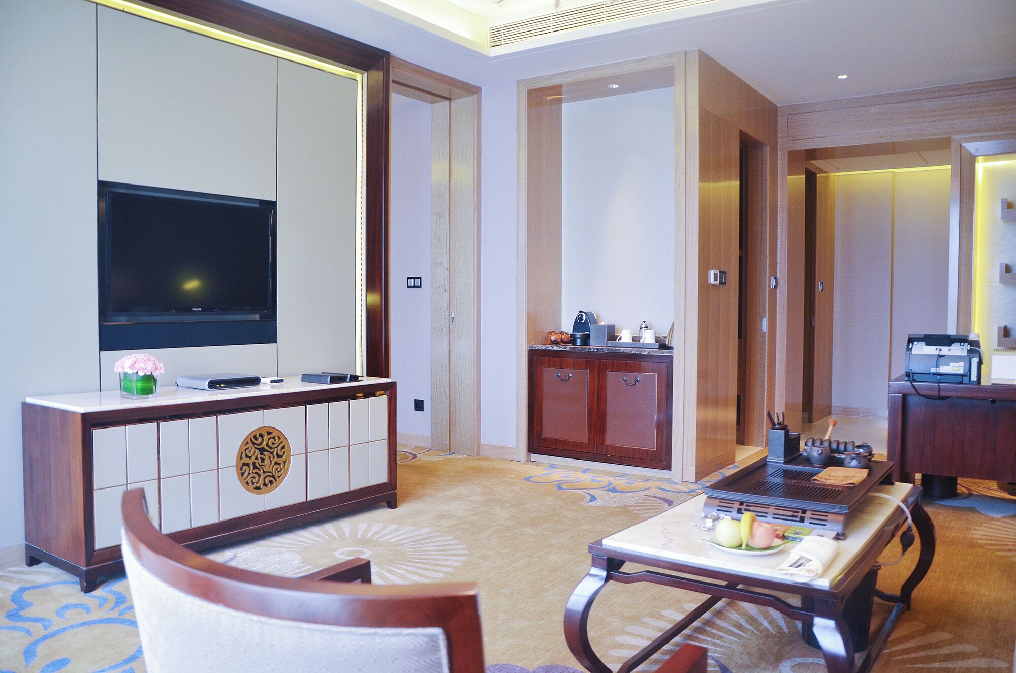 A hotel room with a flat-screen TV, a minibar, a dining table with a laptop, and a sofa.