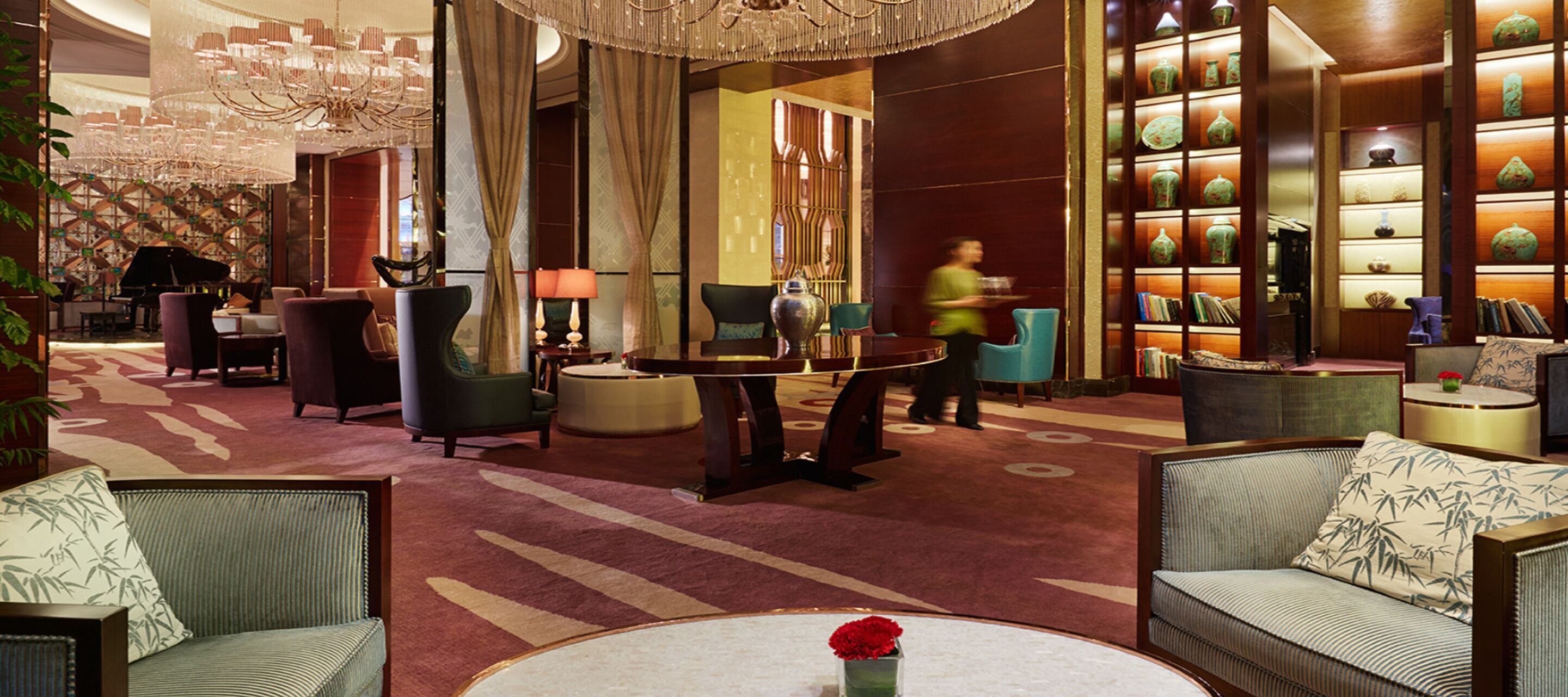 A spacious lobby with a grand chandelier, comfortable seating, and a decorative vase with a red flower on a round table.
