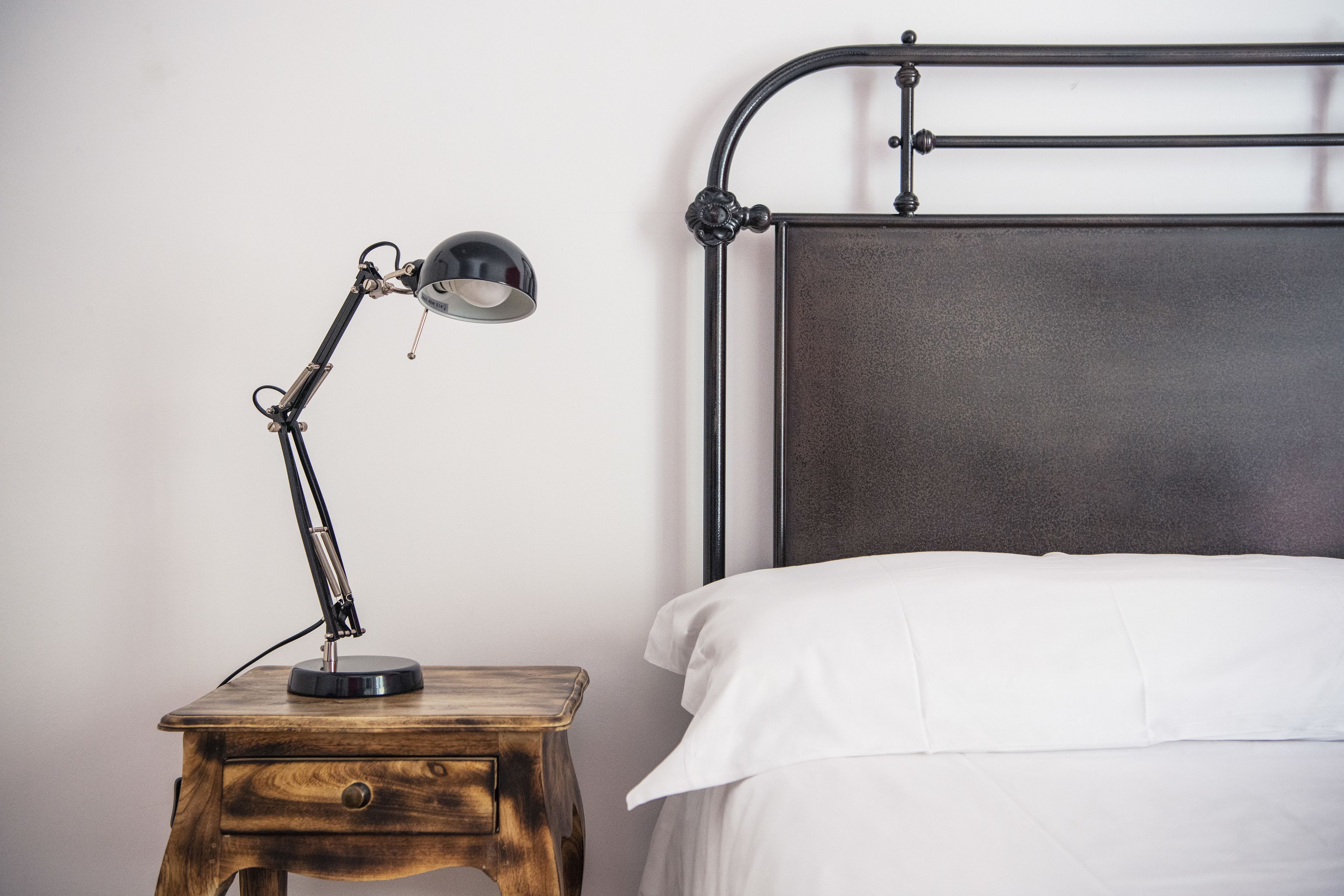 A bed with white bedding, a wooden nightstand with a desk lamp, and a metal headboard.