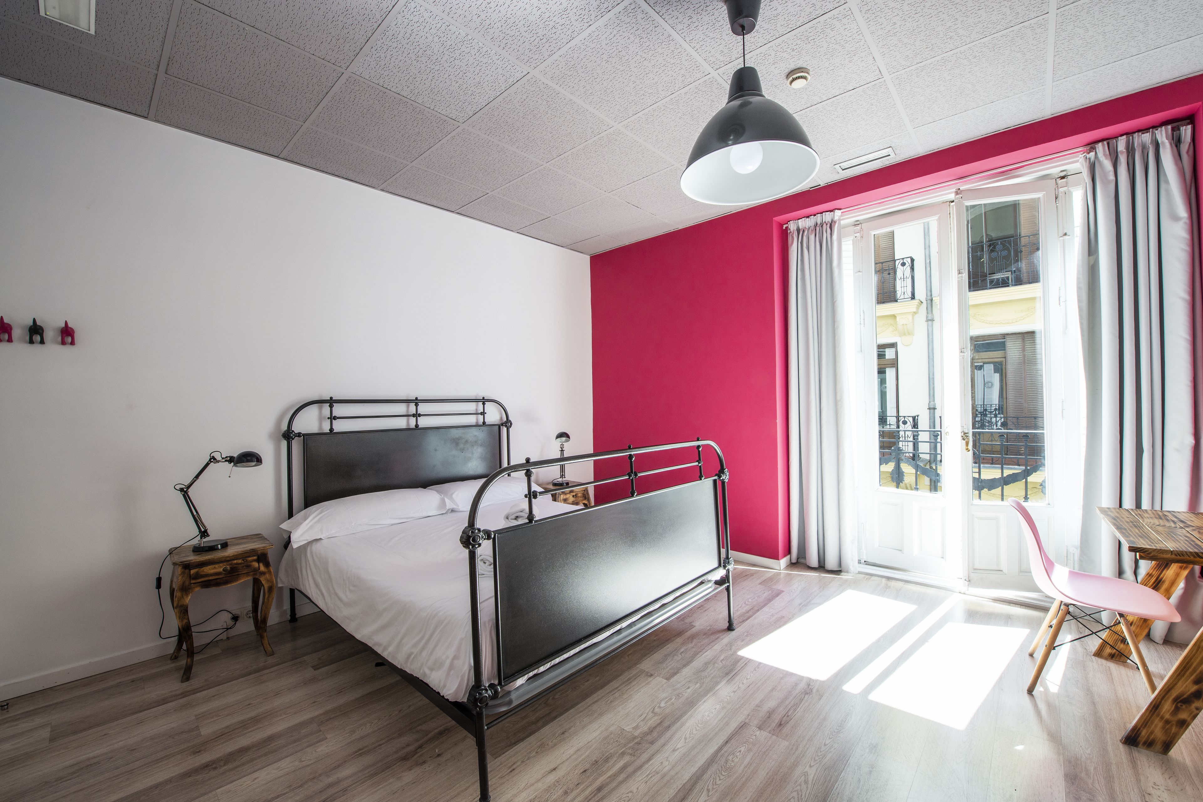 A bedroom with a metal bed frame, a bedside table with a lamp, a pink chair, and a view of a balcony through a sliding door.