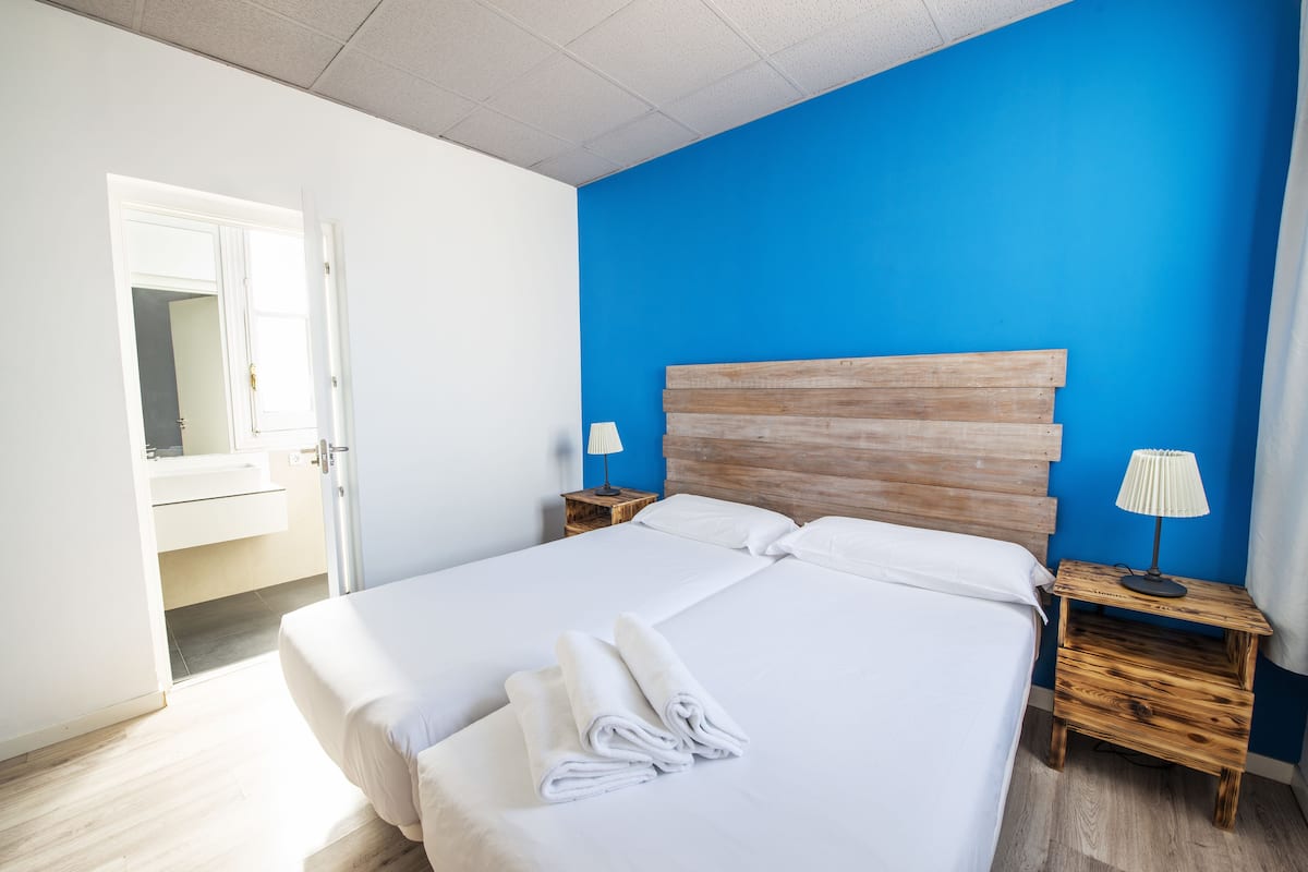 Un lit bien fait, avec du linge de lit et des serviettes blanches, une tête de lit en bois, une lampe de chevet et une petite table de chevet en bois.