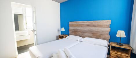 Una cama bien hecha con sábanas y toallas blancas, cabecera de madera, una lámpara de noche y una mesita de noche de madera.