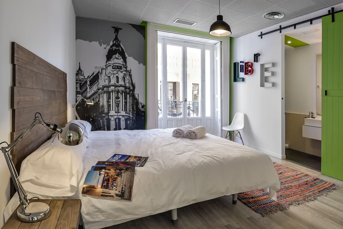 Une chambre moderne avec un grand lit, un bureau avec une lampe, une chaise, un tapis coloré et une porte verte.