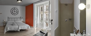 A bedroom with a bed, a bedside table, a lamp, a clock, and a bathroom with a shower and sink. - Safestay Madrid Central (Madrid)