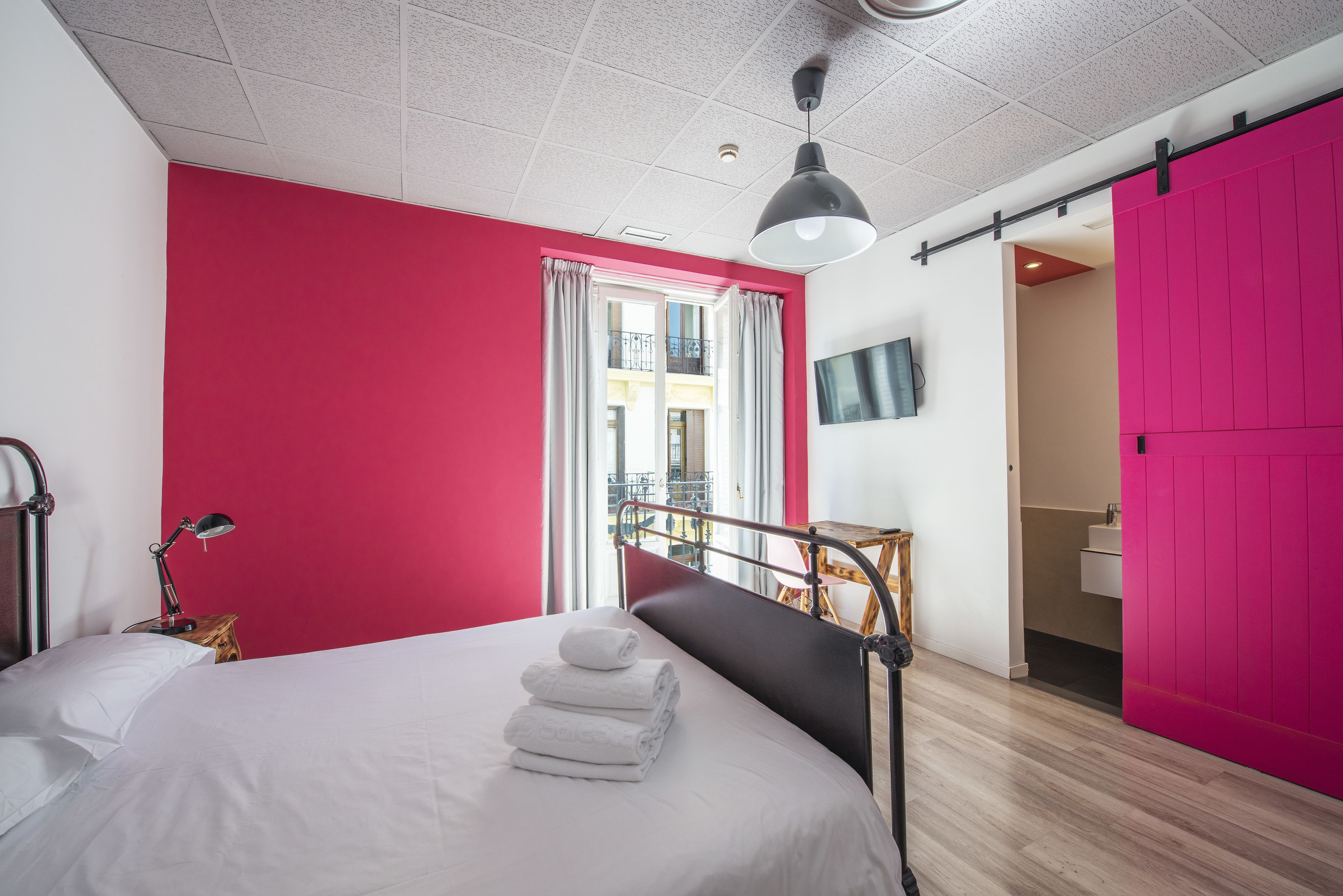 A bedroom with a bed, a bedside lamp, a window with curtains, a pink wall, and a door leading to a bathroom.
