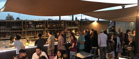 A rooftop bar with outdoor seating, a bar area with shelves of bottles, and a view of the city skyline.