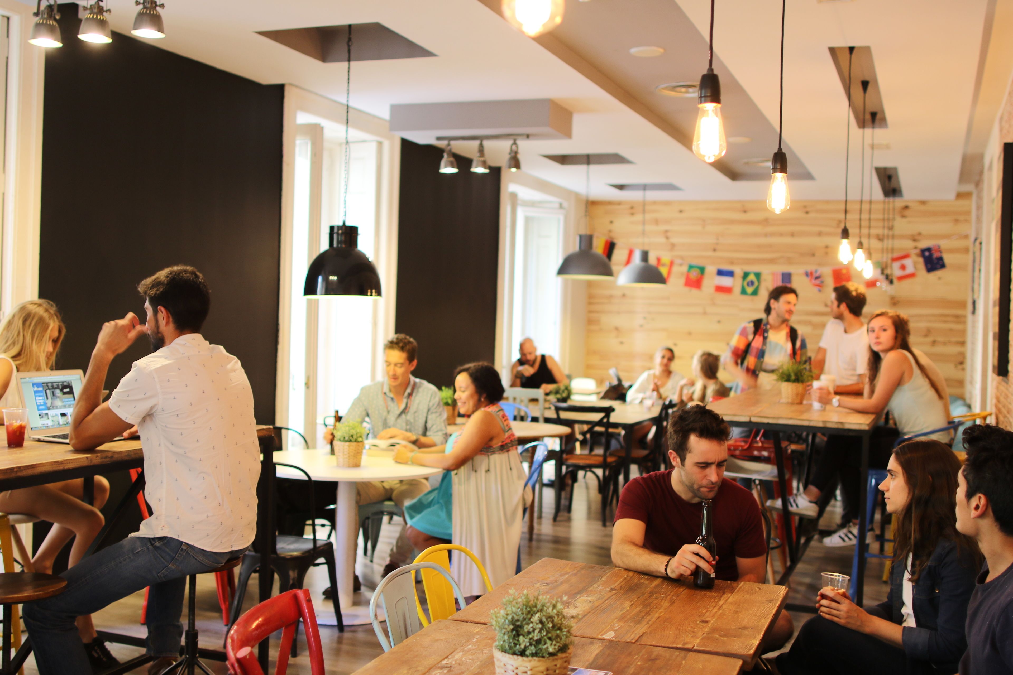 A modern cafe with wooden tables and chairs, people sitting and working, and various flags on the wall.