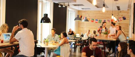 A modern cafe with wooden tables and chairs, people sitting and working, and various flags on the wall.