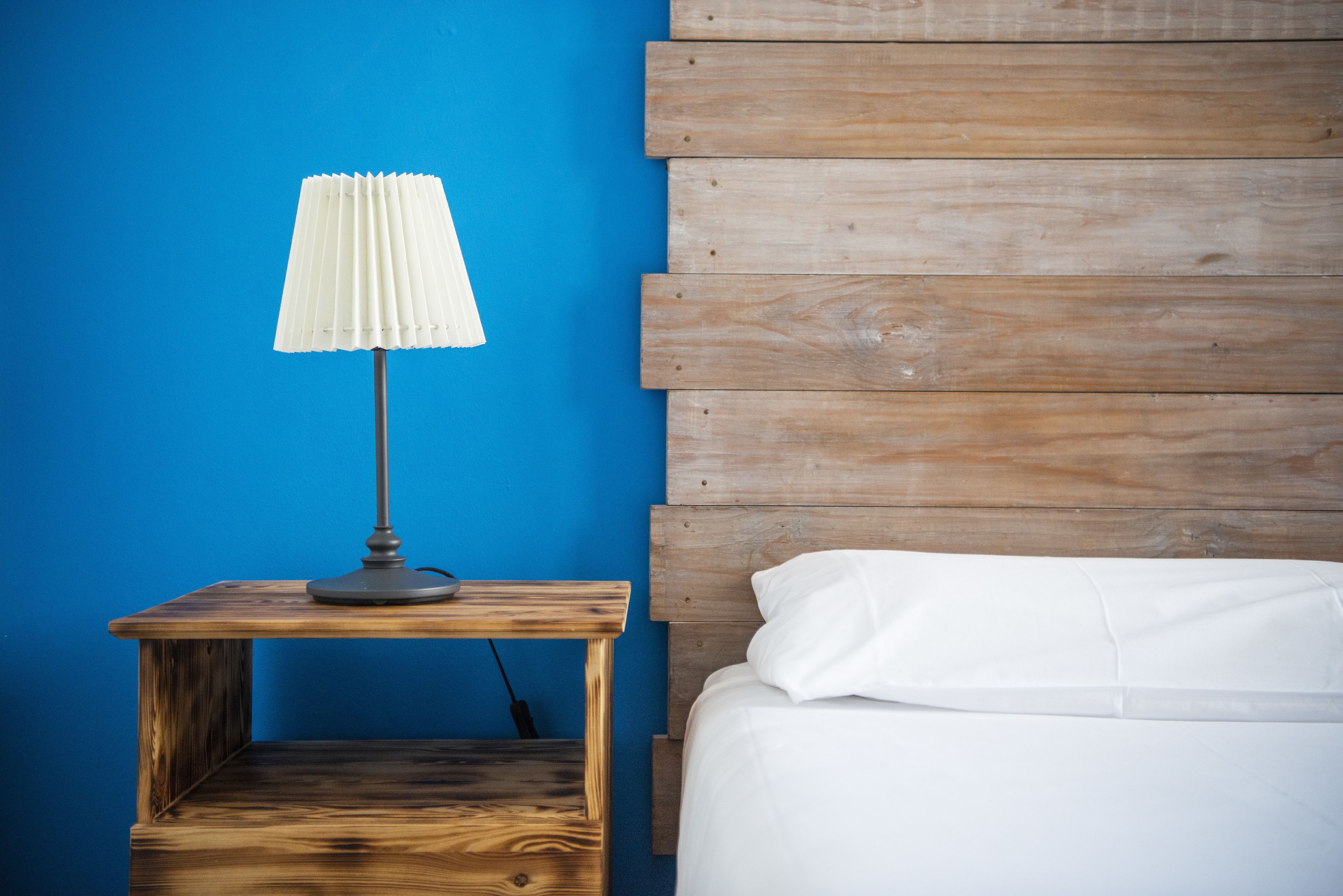 Una cama con ropa de cama blanca, una mesita de noche de madera y una lámpara con pantalla blanca.