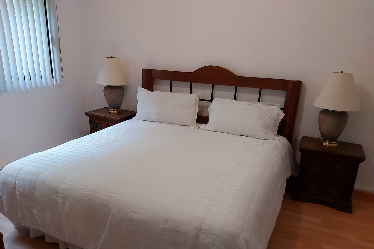 A neatly made bed with a wooden headboard, two bedside tables with lamps, and a window with blinds.