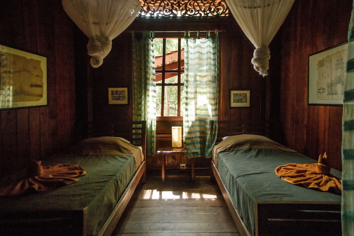 Une chambre avec deux lits simples, un plancher en bois, une fenêtre avec des rideaux et des tableaux encadrés accrochés aux murs.