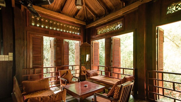 A wooden interior with a table and chairs, large windows with shutters, and a view of greenery outside. - Rajabori Villas (Kratie)