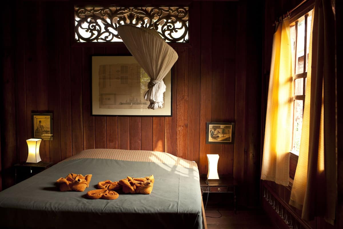 Une pièce avec un lit, une fenêtre avec des rideaux, une lampe fixée au mur et un tableau encadré.