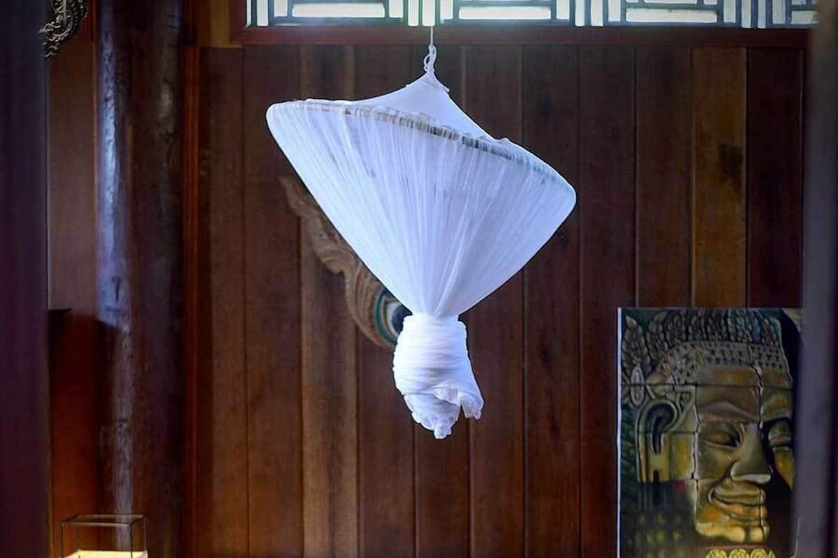 Un lit recouvert d’une couvre-lit verte et surmonté d’un dais blanc, des murs en bois et un tableau représentant Bouddha.