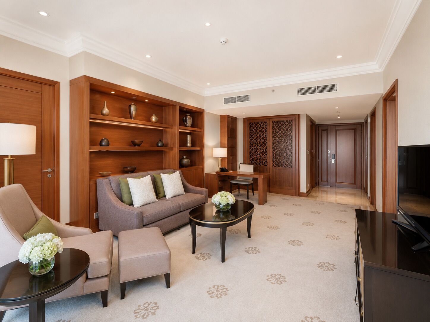 A modern living room with a sofa, armchair, ottoman, coffee table, and a TV. There are wooden shelves with decorative items, a patterned carpet, and a decorative screen.