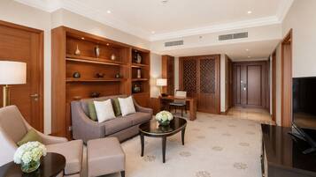 A modern living room with a sofa, armchair, ottoman, coffee table, and a TV. There are wooden shelves with decorative items, a patterned carpet, and a decorative screen.