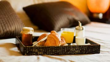 Bandeja de desayuno con croissants, zumo de naranja y una botella de miel sobre una cama con almohadas y una lámpara al fondo.