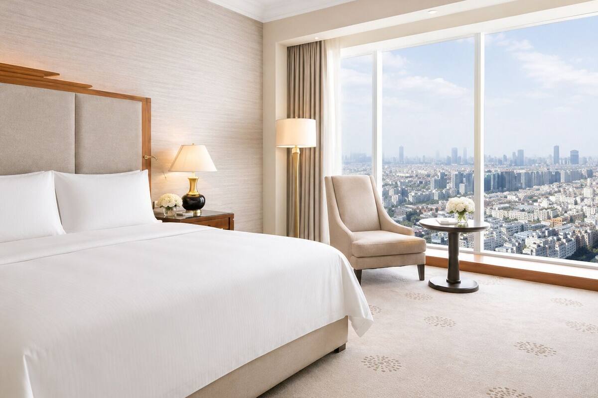 A hotel room with a large bed, a chair, a small table, and a view of a cityscape through a large window.