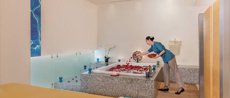 Un cuarto de spa con una bañera, una mesa de masajes y una persona disponiendo pétalos de rosa.