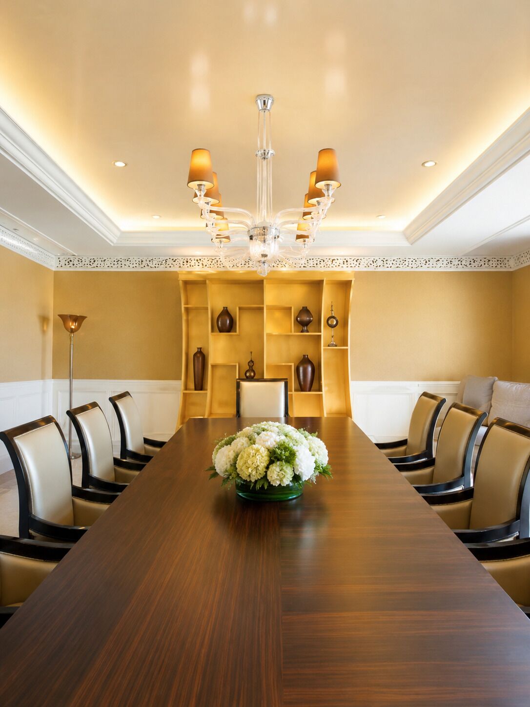 A conference room with a long wooden table, chairs, a chandelier, and shelves with decorative items.