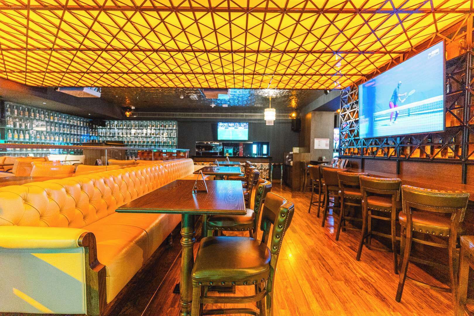 A bar area with a long counter, leather seating, and a large screen displaying a tennis match.
