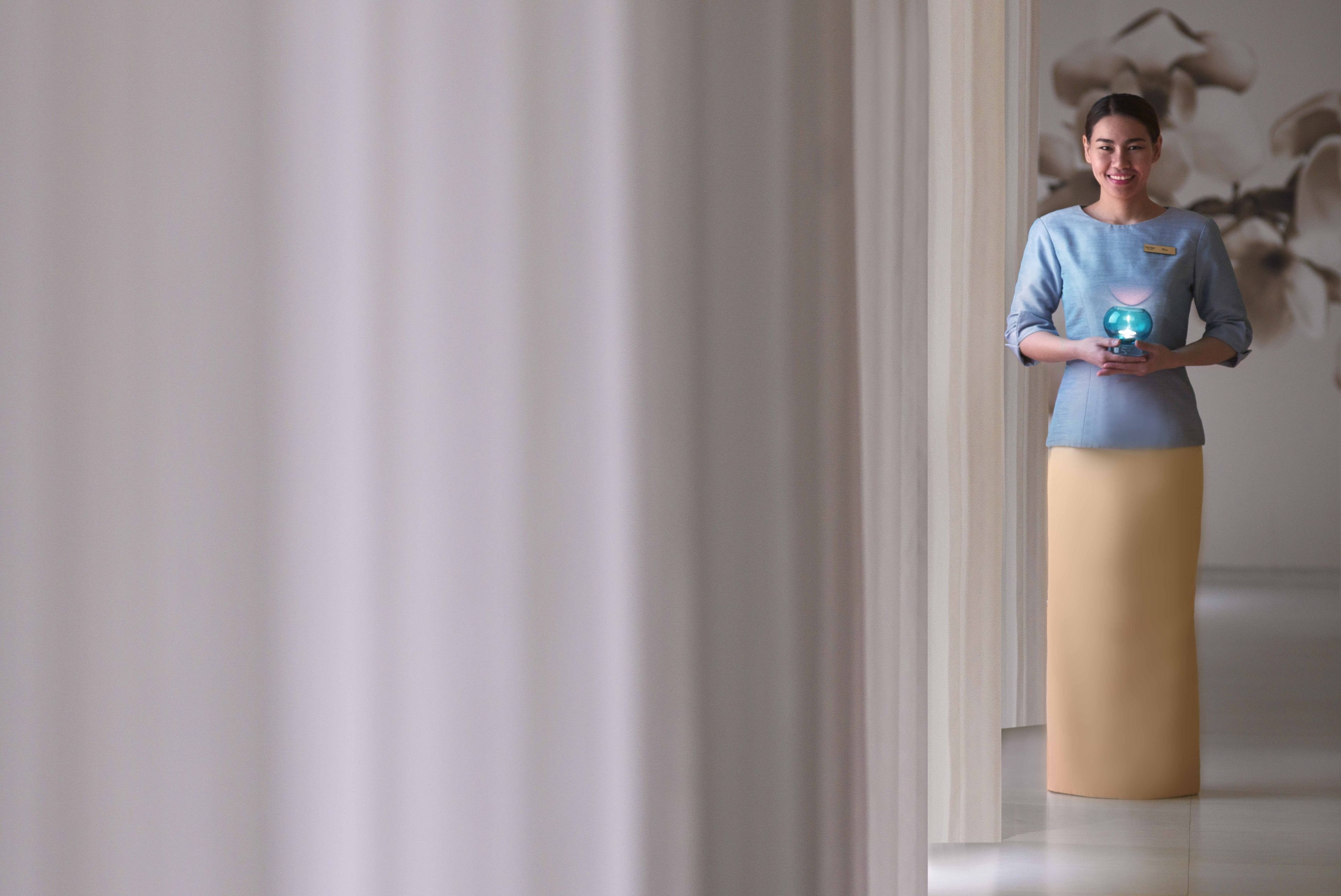 A person standing in a hallway with a light-colored floor and walls, holding a blue object.