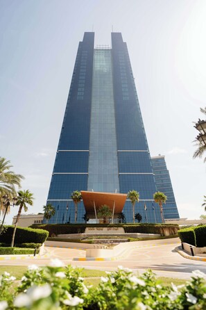A tall, modern skyscraper with a glass facade, surrounded by well-maintained greenery and a landscaped area with a curved pathway.
