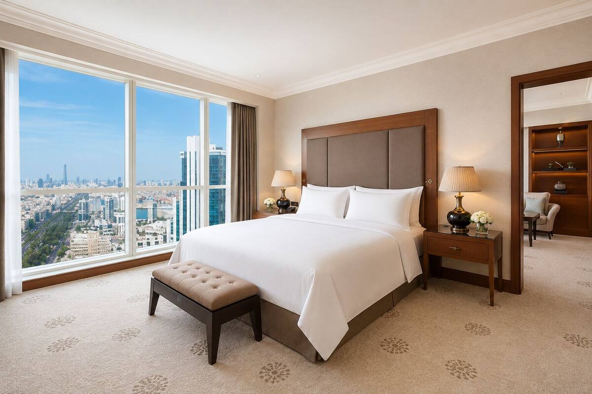 A hotel room with a large bed, a nightstand, a lamp, a chair, and a view of the city.