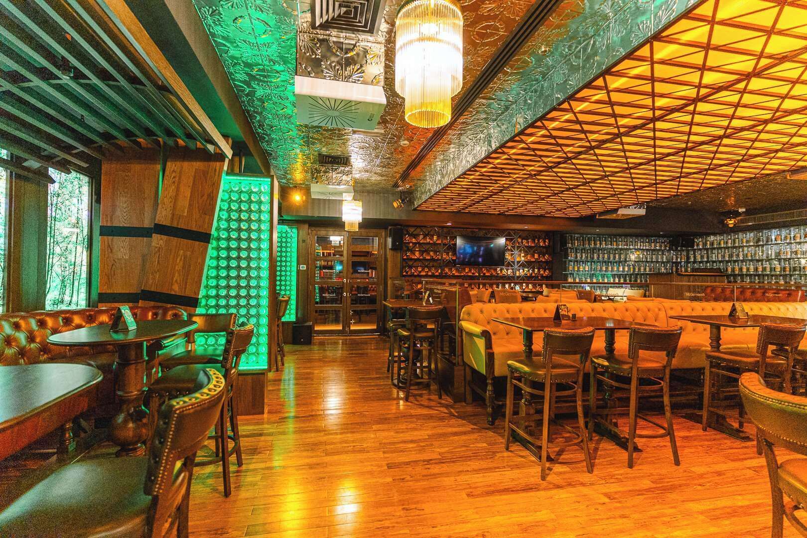 Eine Bar mit Holzboden, einer beleuchteten grünen Wand und verschiedenen Sitzgelegenheiten, darunter Tische und Stühle.