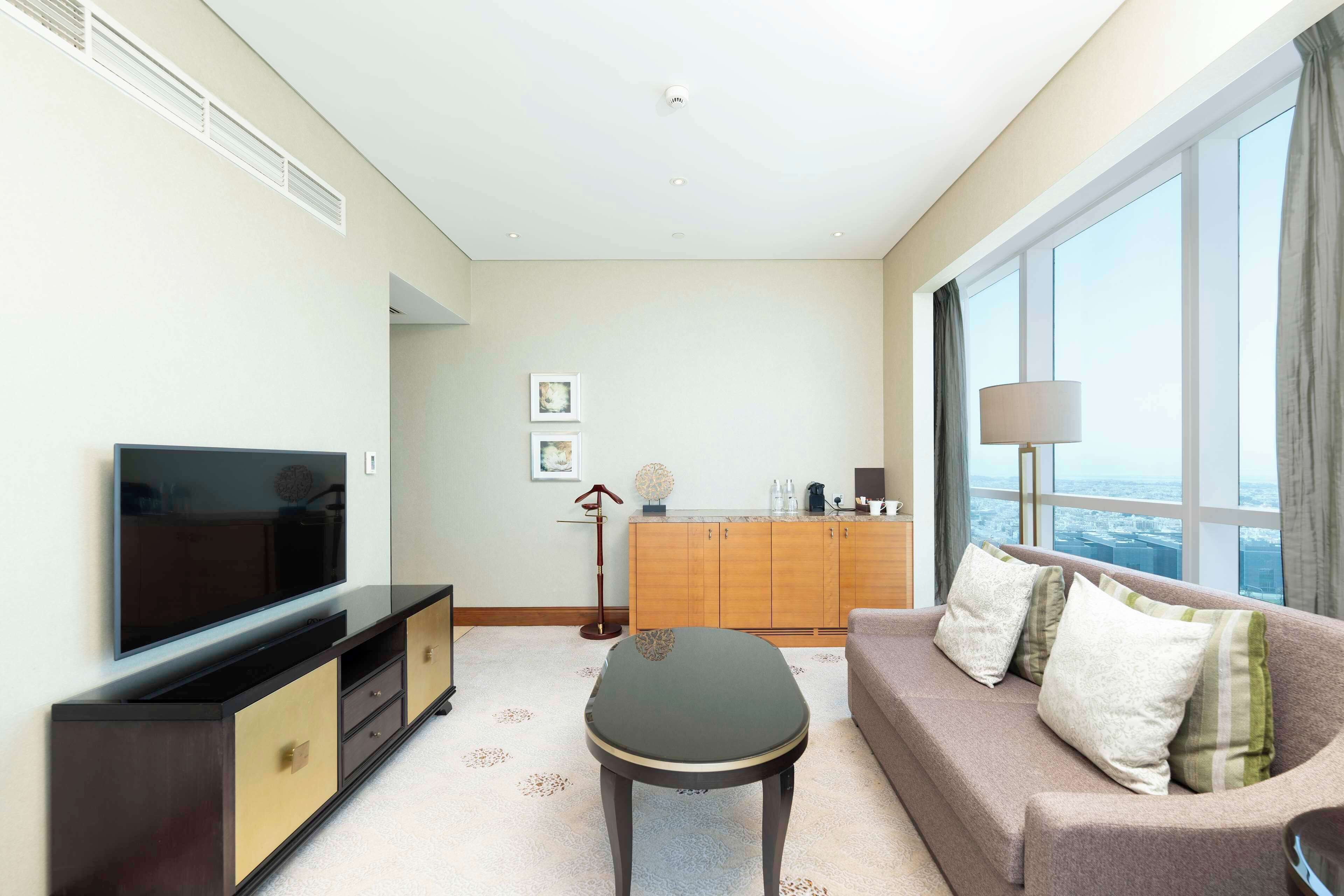 A modern living room with a flat-screen TV, a sofa with cushions, a coffee table, and a large window offering a city view.