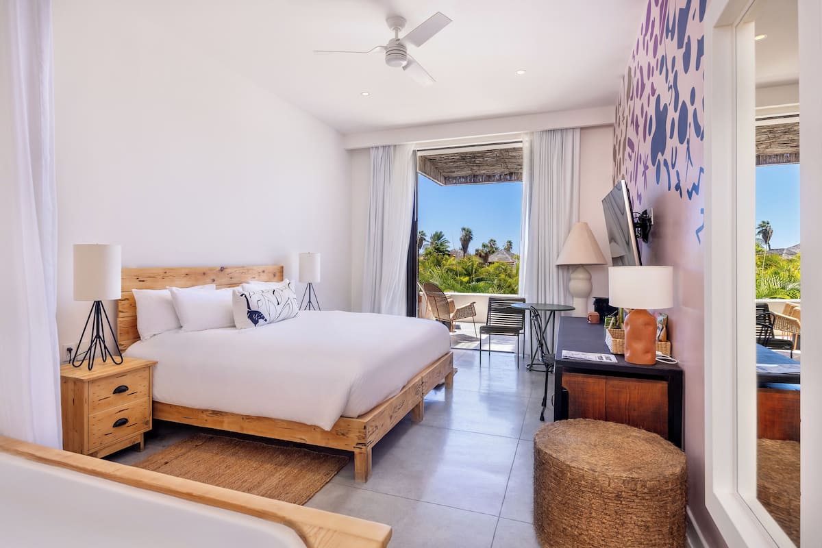 A modern hotel room with a wooden bed, a desk, and a balcony with a view of palm trees.