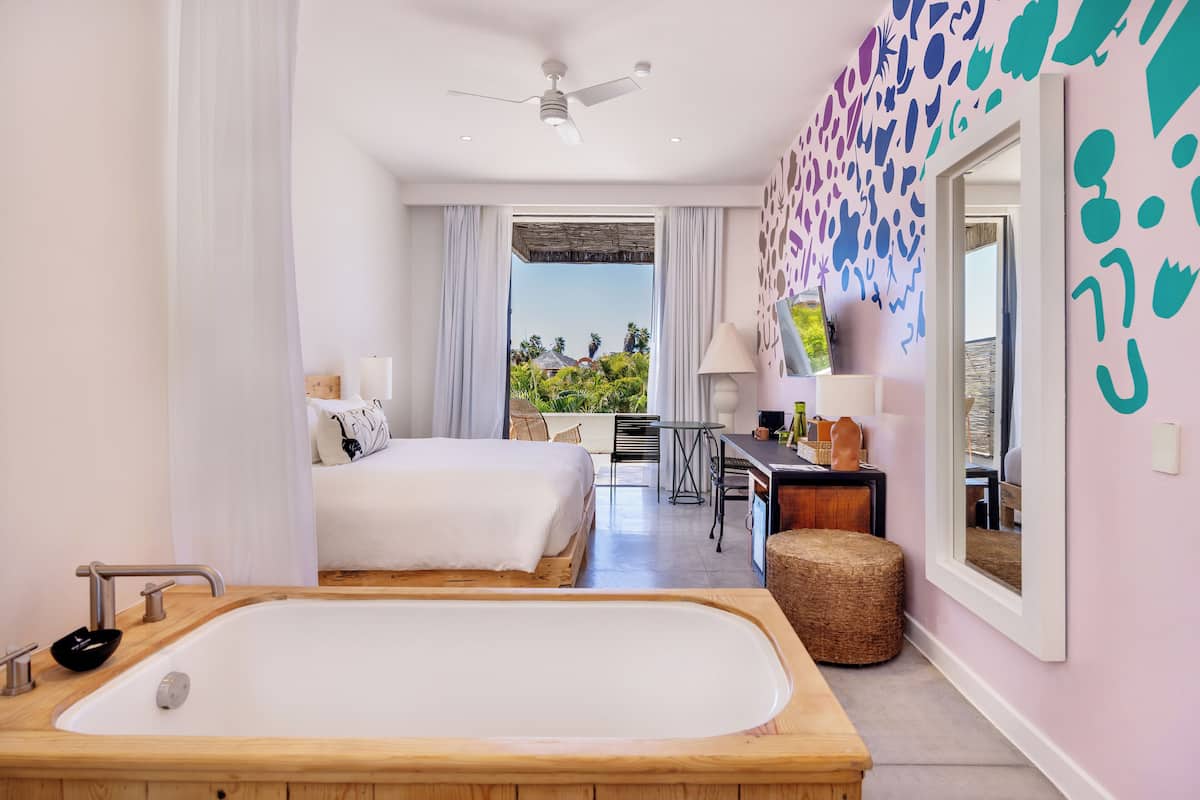 A modern hotel room with a large bed, a wooden bathtub, a desk with a chair, and a balcony with a view of greenery.