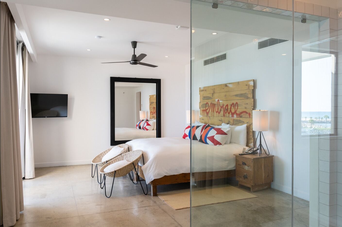A modern bedroom with a bed, a television, a ceiling fan, and a view of the outdoors.