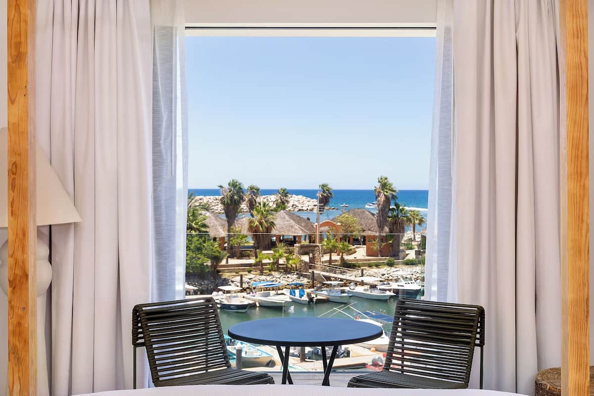 A hotel room with a large window, a bed, a table with chairs, and a view of a beach and boats.