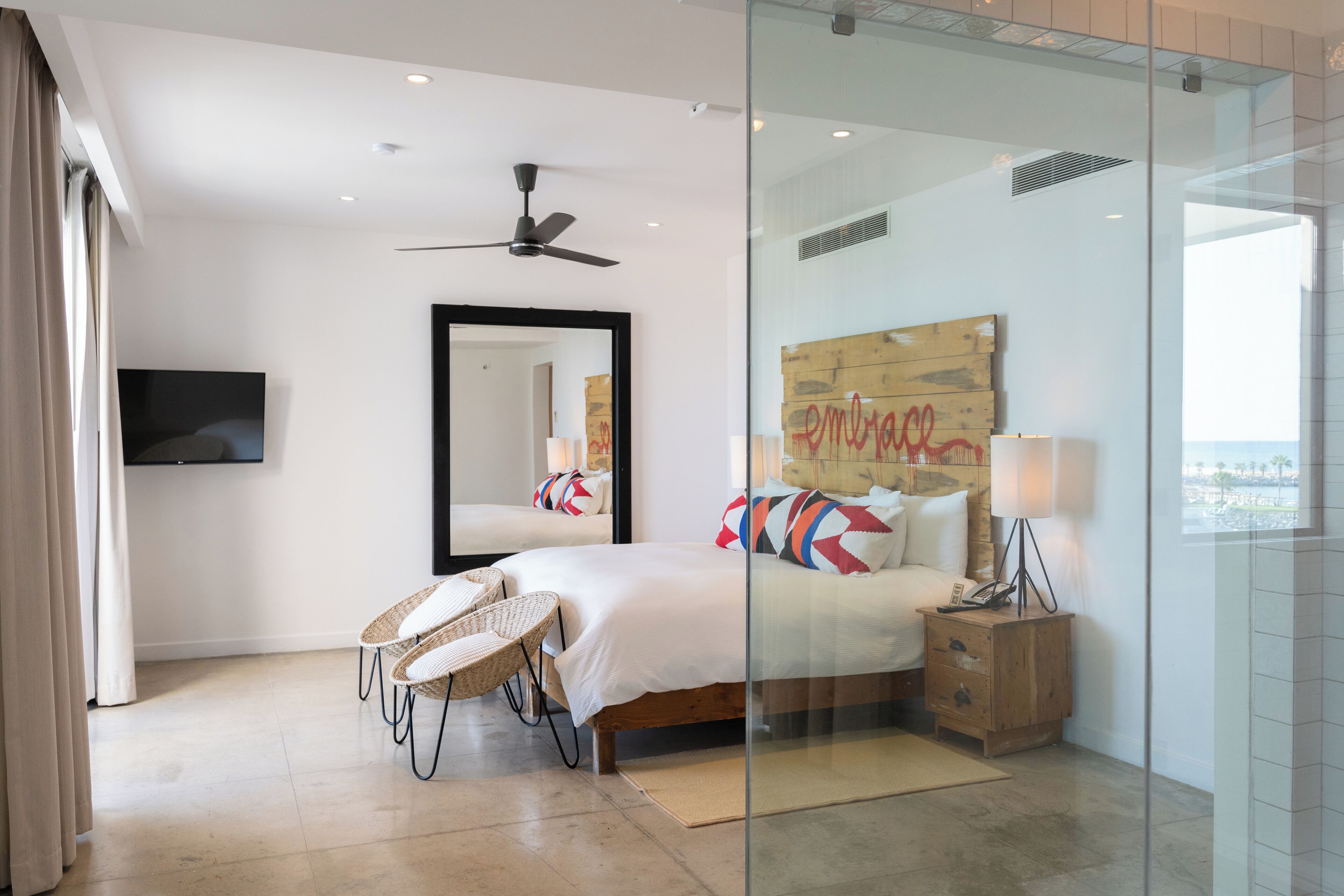 A modern hotel room with a large bed, a ceiling fan, a flat-screen TV, wicker chairs, a wooden bedside table with a lamp, and a large window offering a view.