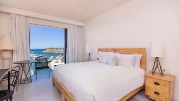 Ein Schlafzimmer mit einem großen Bett, einem hölzernen Kopfteil, einem Nachttisch, einem Tisch und einem Stuhl. Durch ein großes Fenster ist Meer und Bebauung zu sehen.