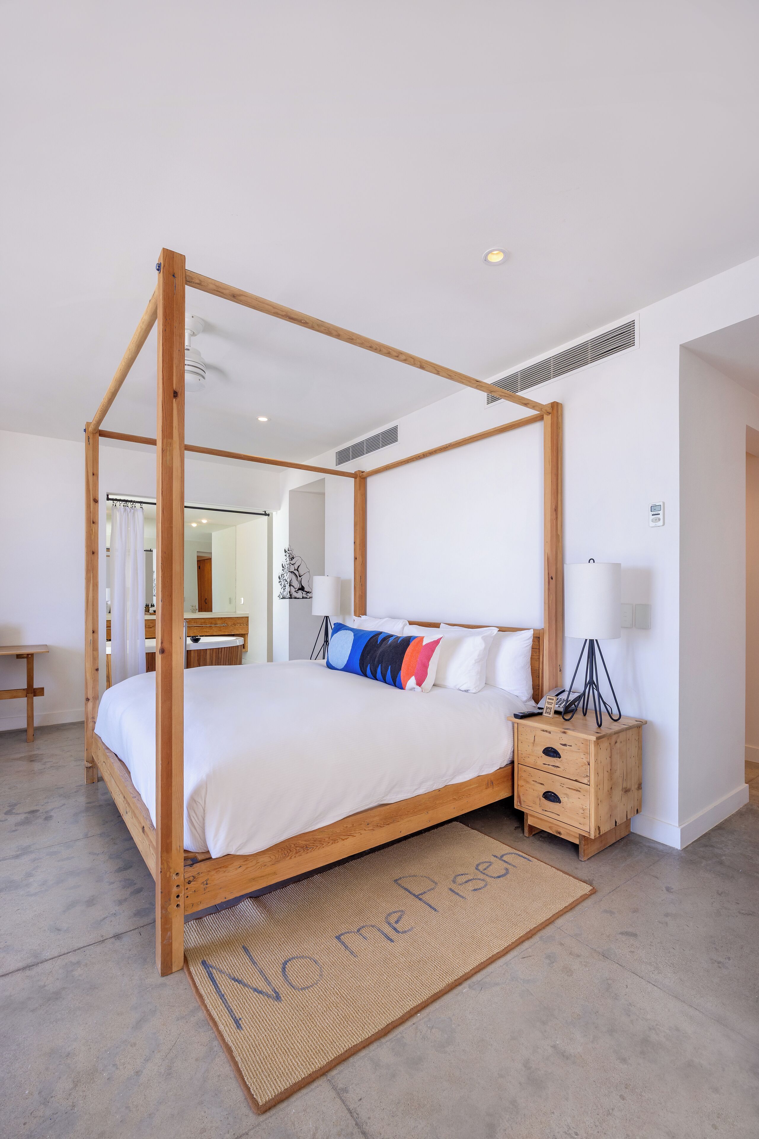 Uma cama com dossel e estrutura de madeira, uma mesinha de cabeceira com abajur e um tapete de entrada com texto.
