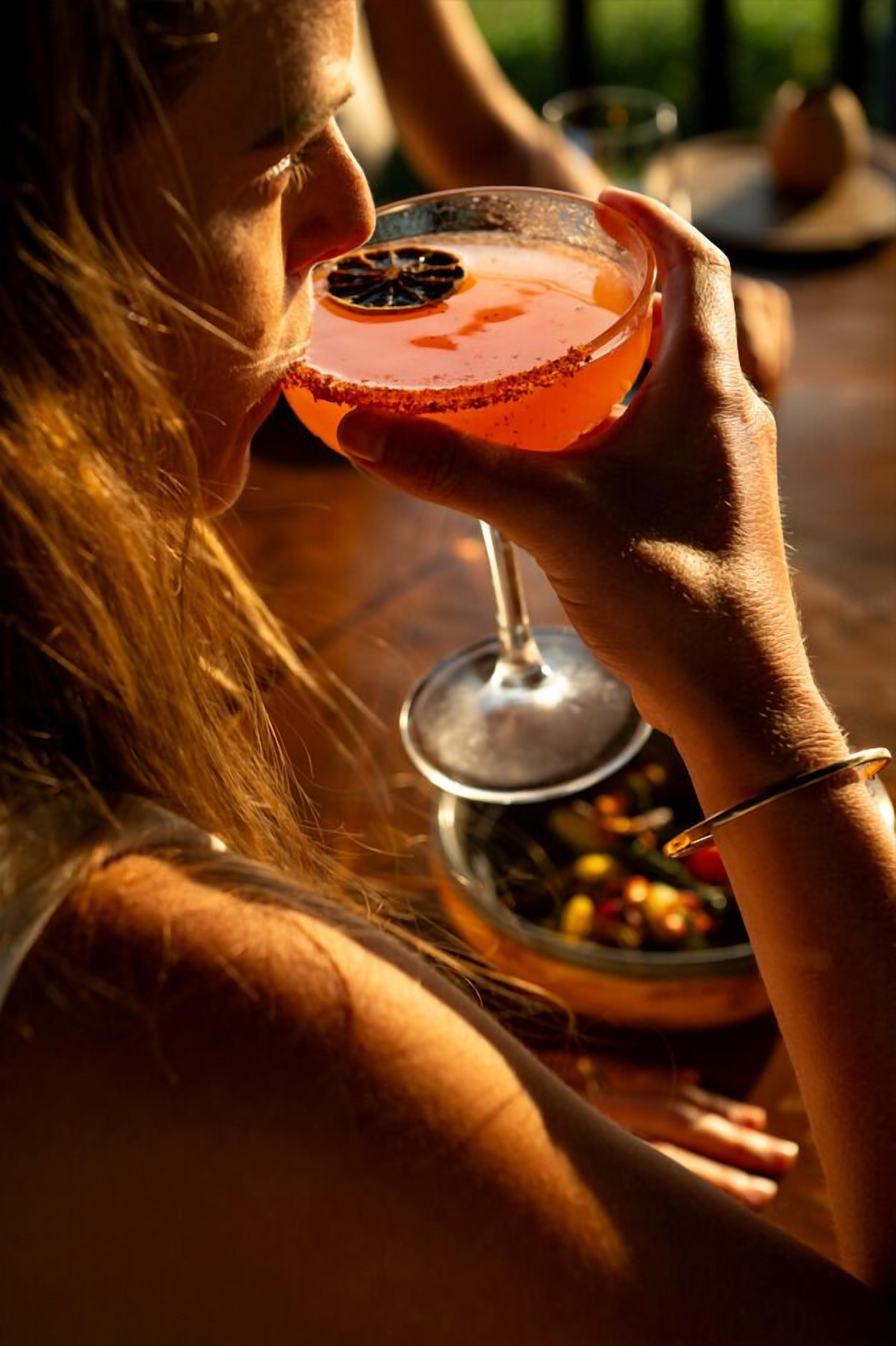 A person holding a cocktail with a salted rim, a glass of water, and a bowl of fruit on a table.