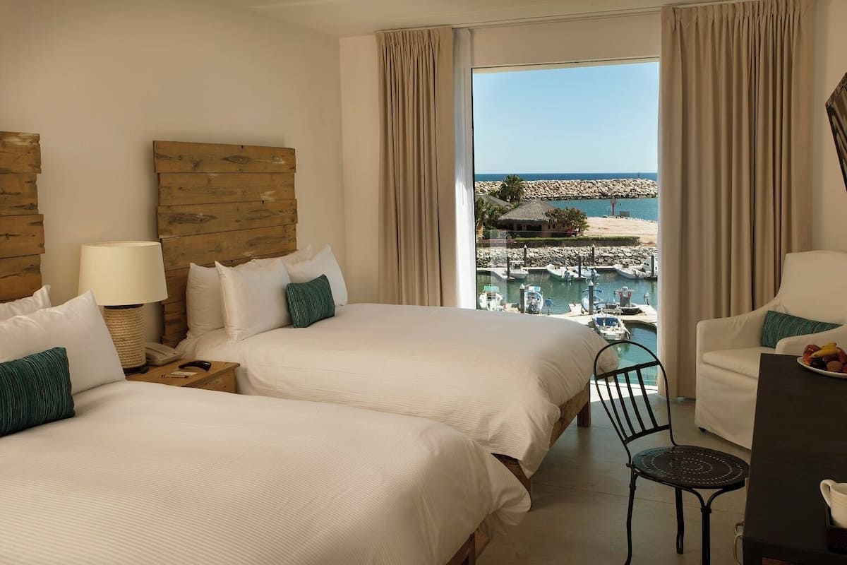 A hotel room with two beds, a desk, a chair, and a view of the sea and marina.
